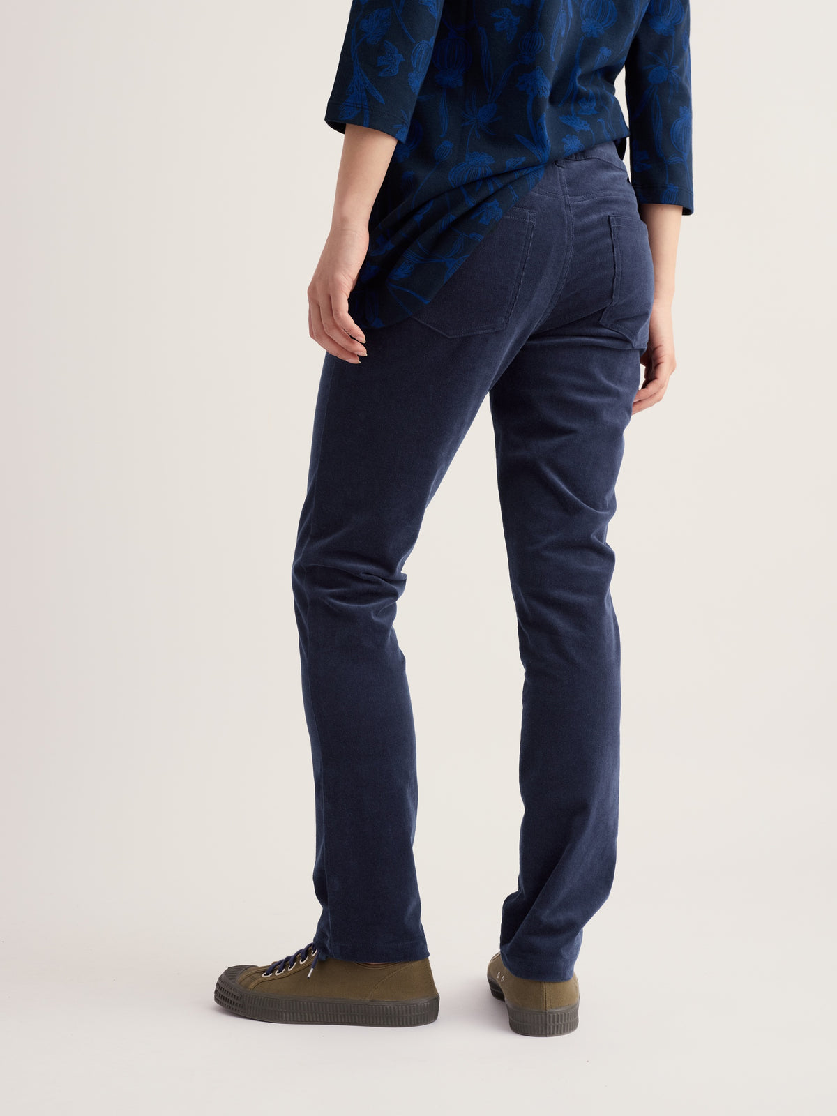 Rear view of a woman wearing navy corduroy trousers and a patterned blue top against a plain background.