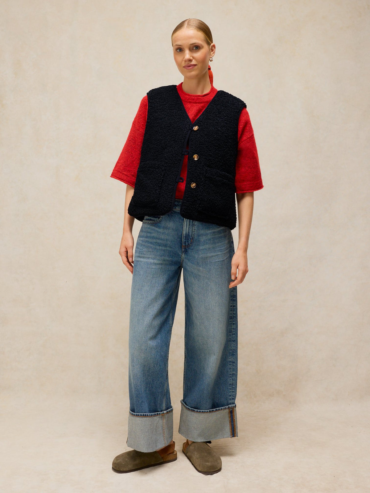 A woman modeling a navy reversible gilet over a red top and wide-leg cuffed jeans against a neutral studio background.