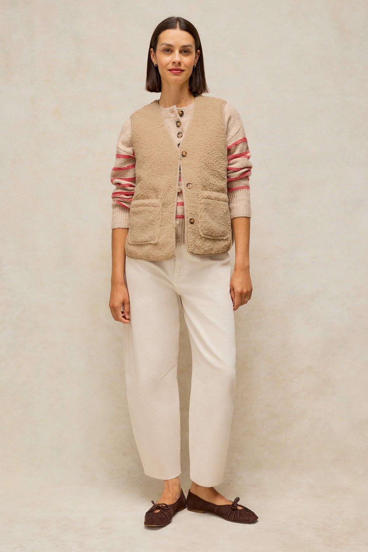 Woman wearing a beige buttoned gilet over a striped sweater and light trousers.
