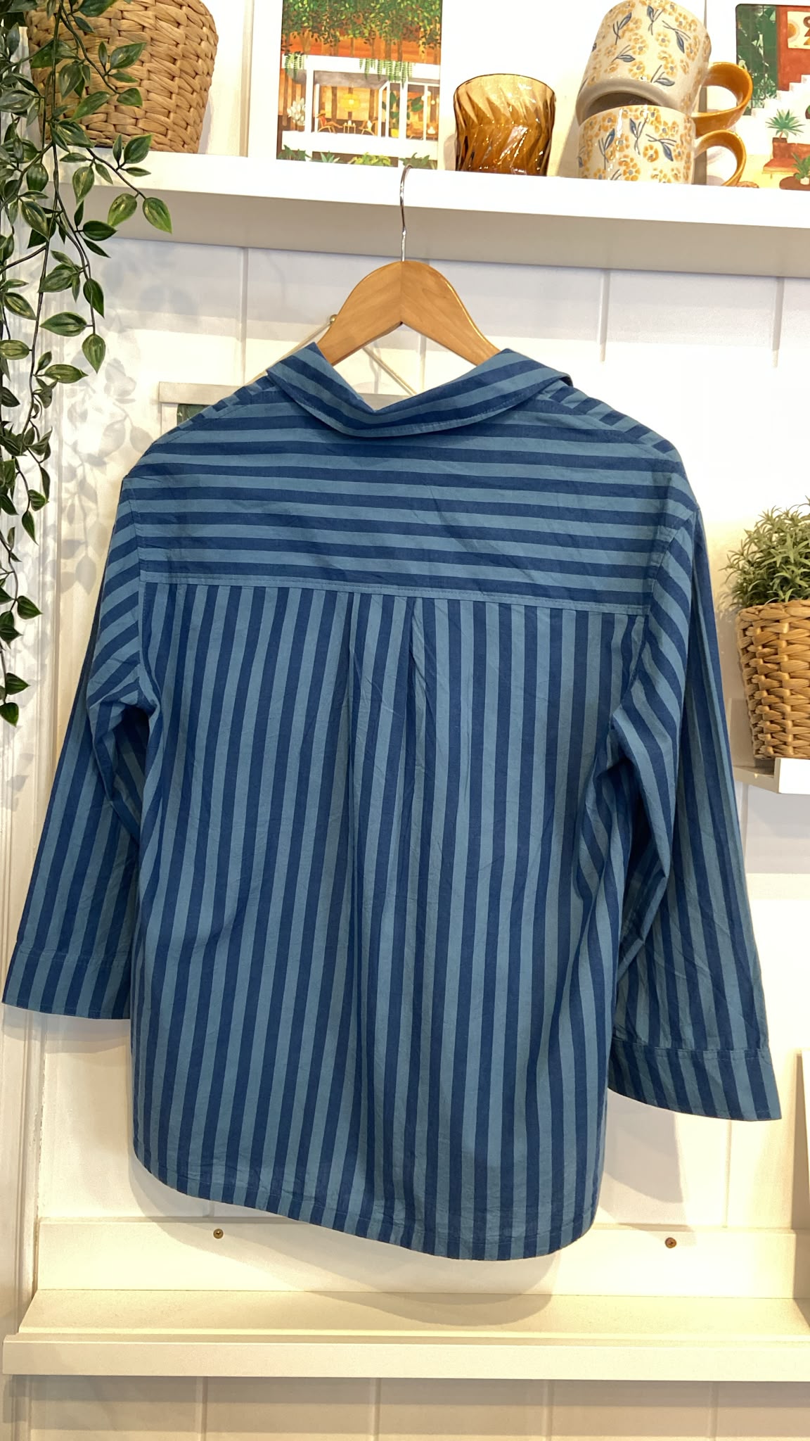 Rear view of a blue striped Seasalt Cornwall cotton shirt hanging on a wooden hanger against a white wall.