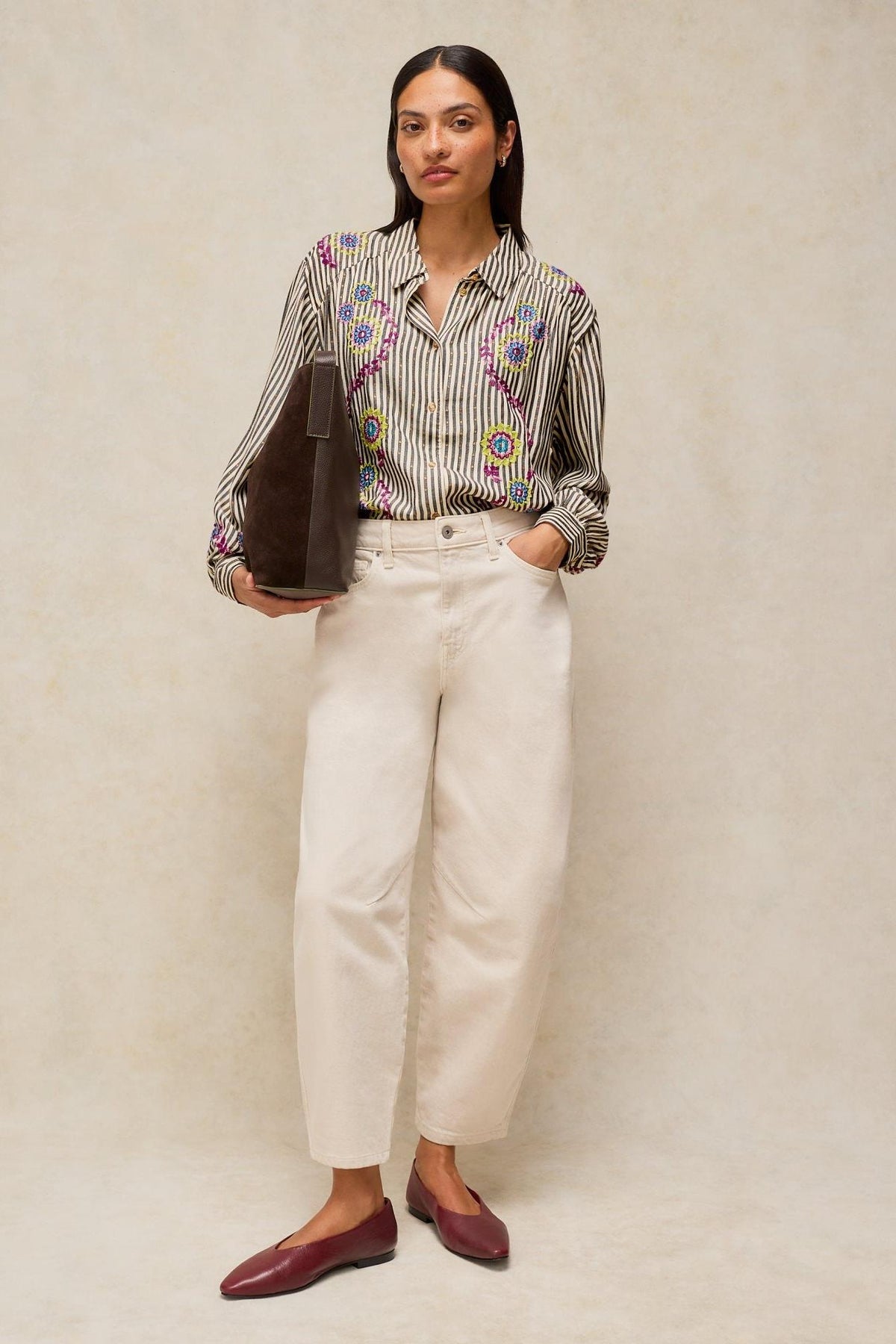 A woman wearing a White Stuff striped blouse with colorful embroidered patterns and cream-colored trousers.