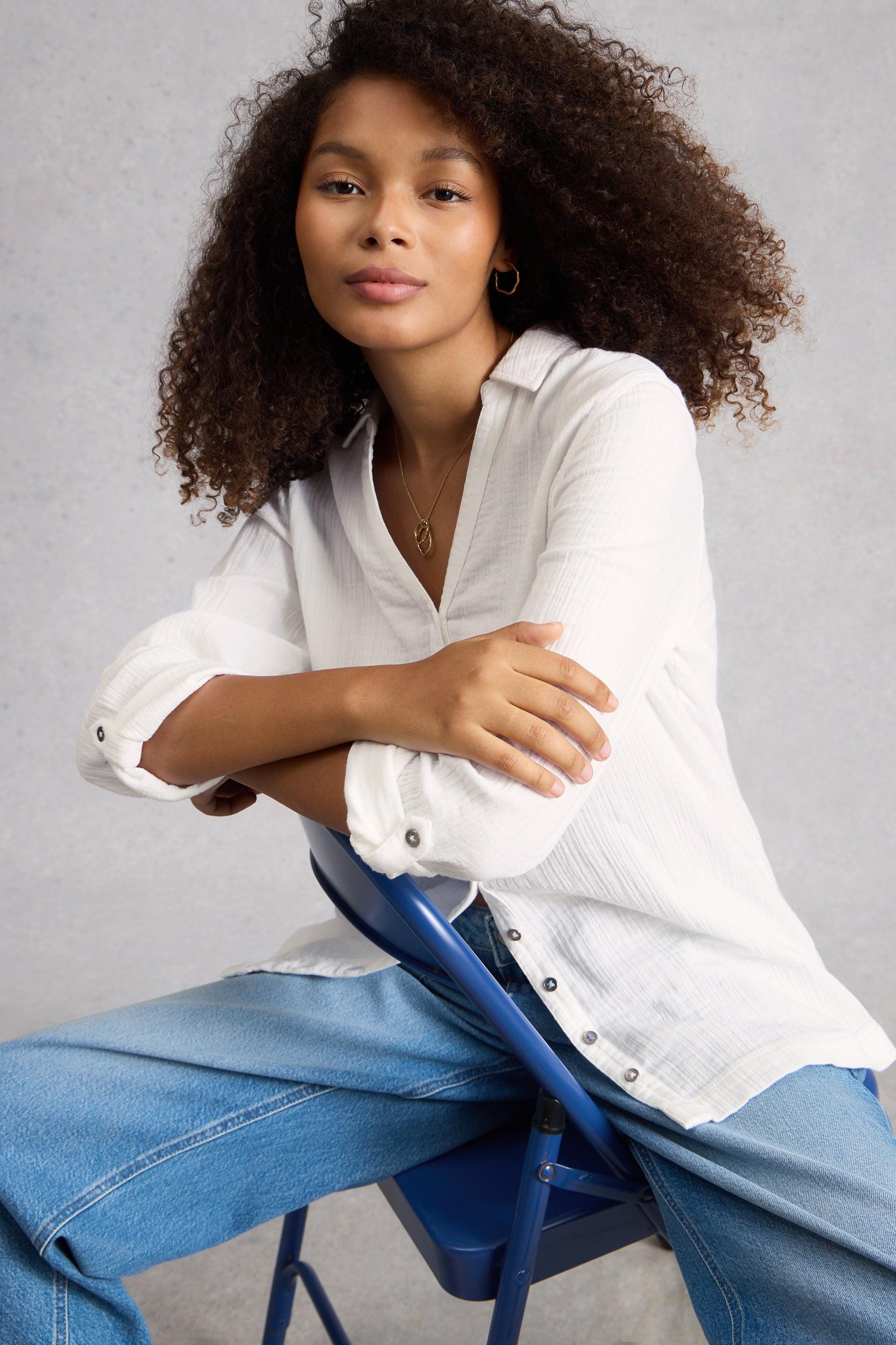 Woman wearing a white organic cotton shirt and blue jeans sitting on a blue chair against a gray background