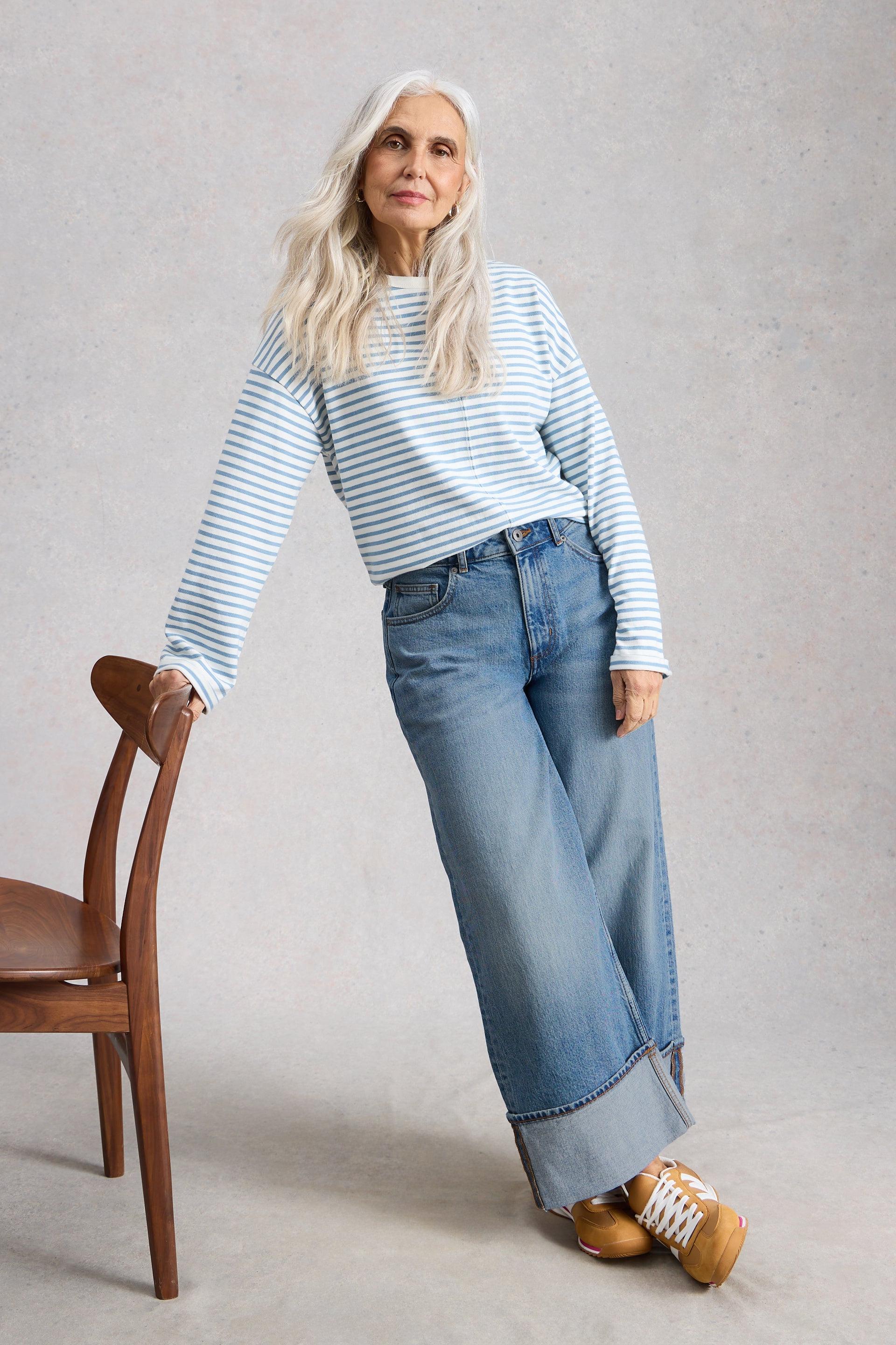 Woman wearing a striped sweater and straight jeans standing next to a wooden chair against a plain background