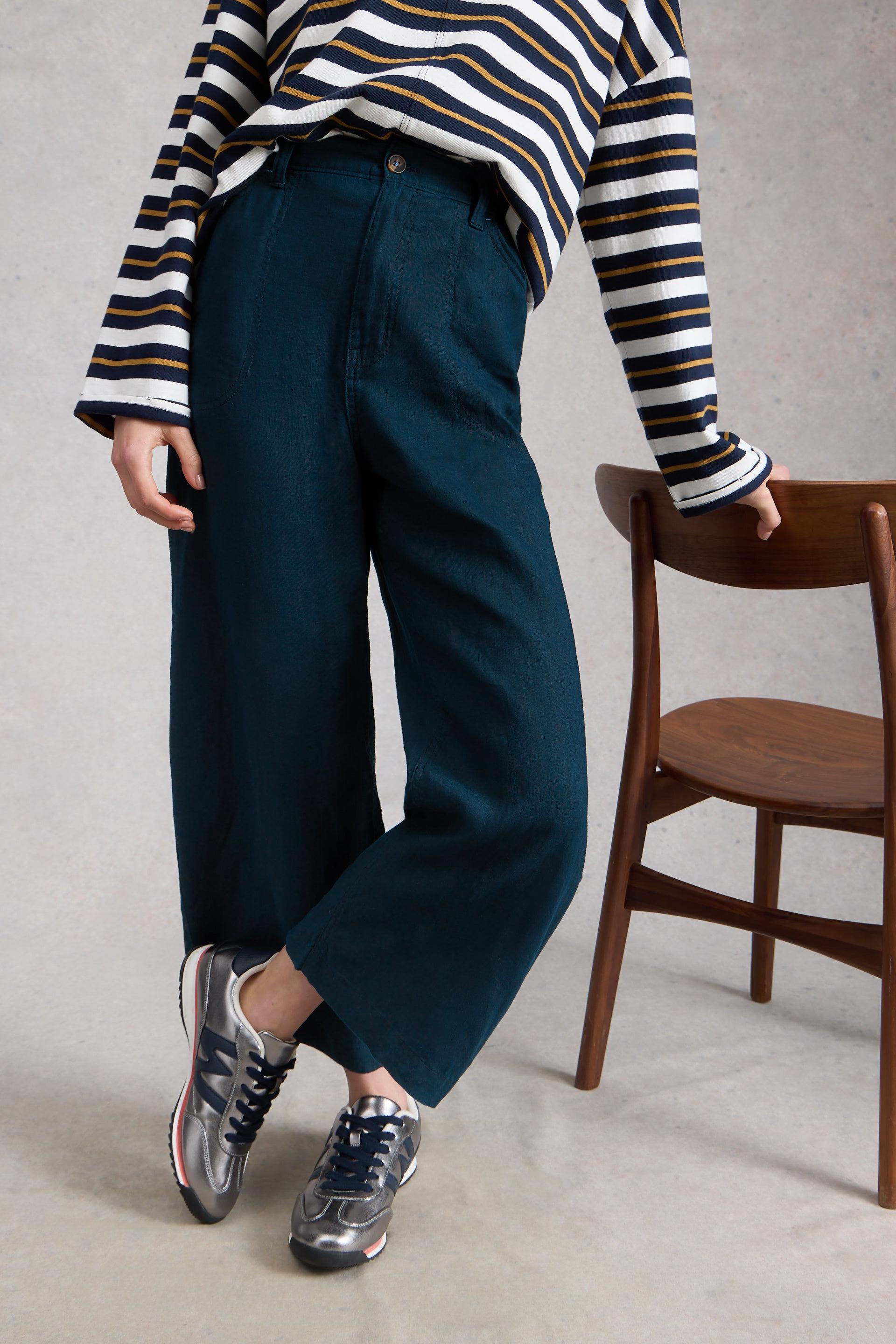 Person wearing a striped shirt and navy wide leg trousers standing next to a wooden chair.