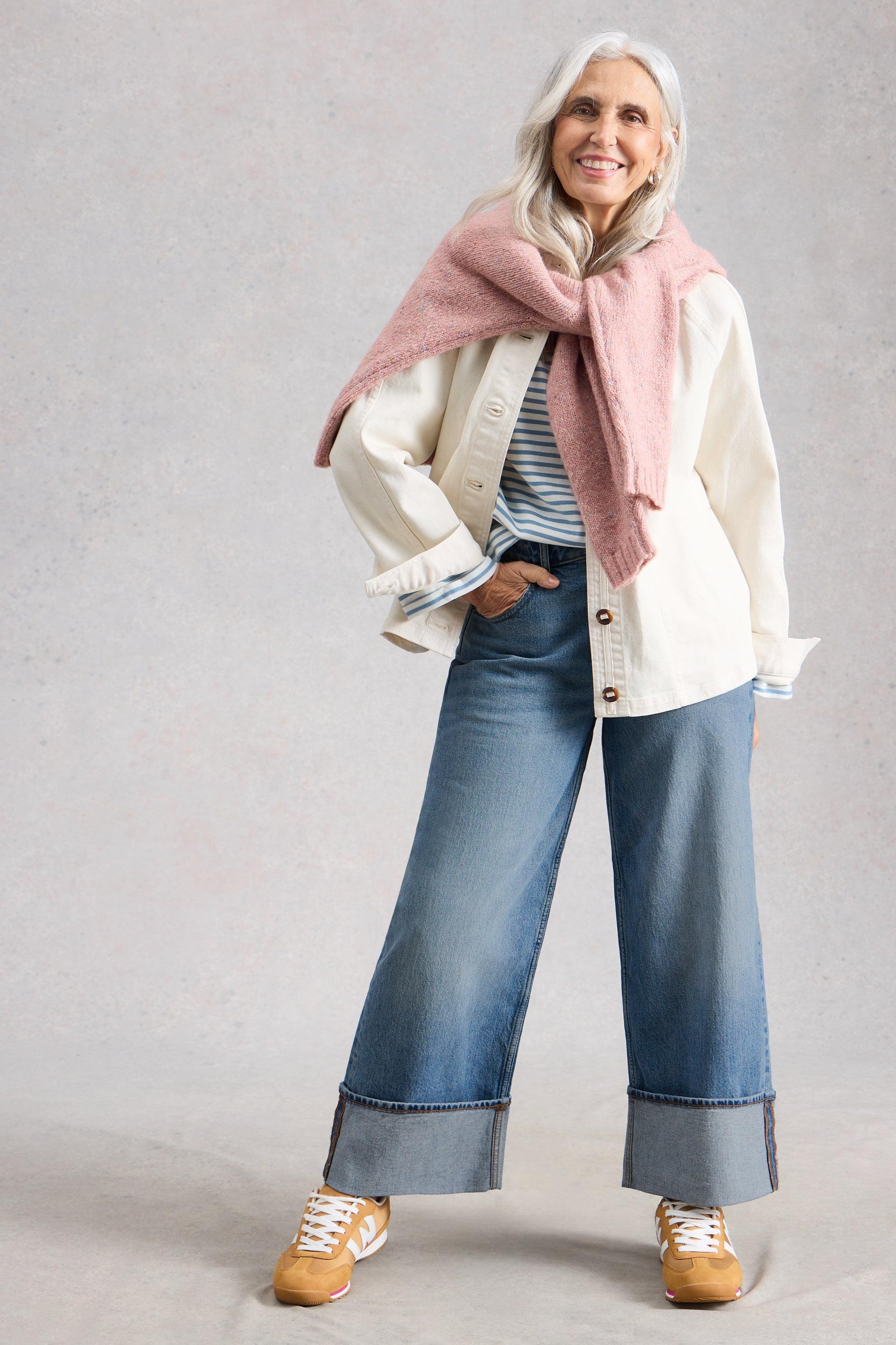 Woman wearing a white jacket, striped shirt, pink scarf, and blue wide leg cropped jeans on a gray background