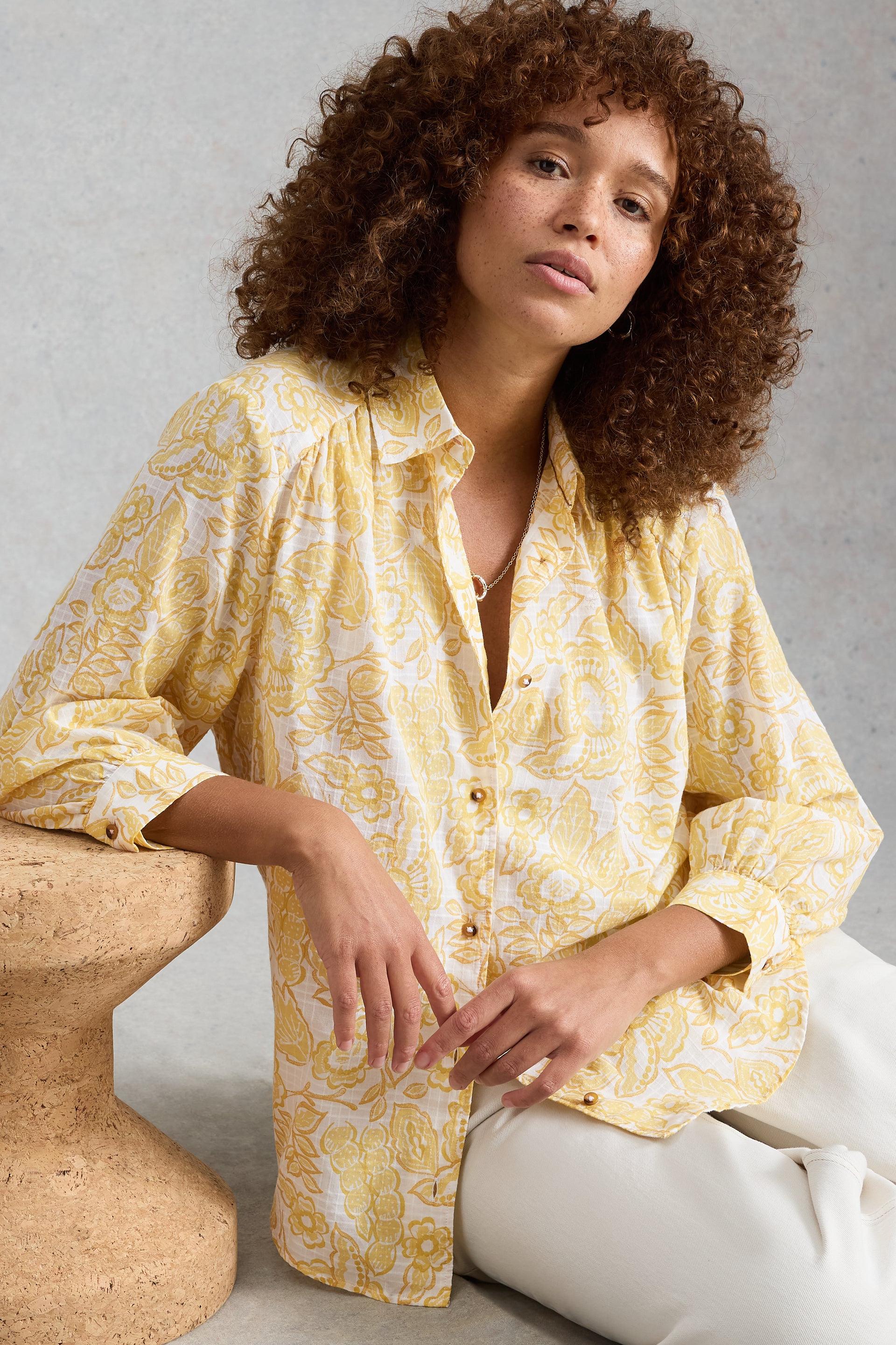 Woman wearing a yellow patterned blouse sitting on the floor against a gray background