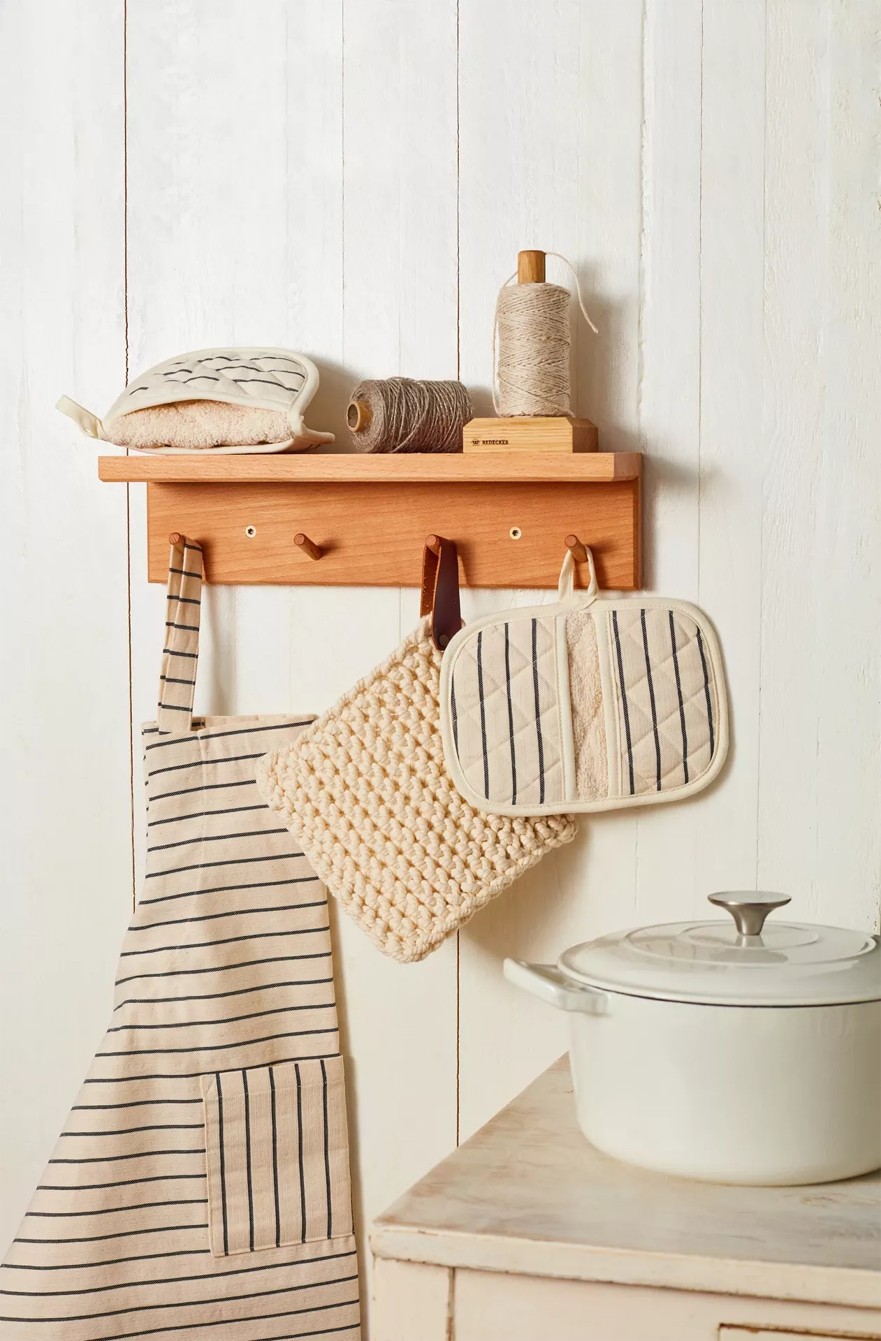 Wooden shelf with kitchen items including a pot, apron, and oven mitt on a white wall.