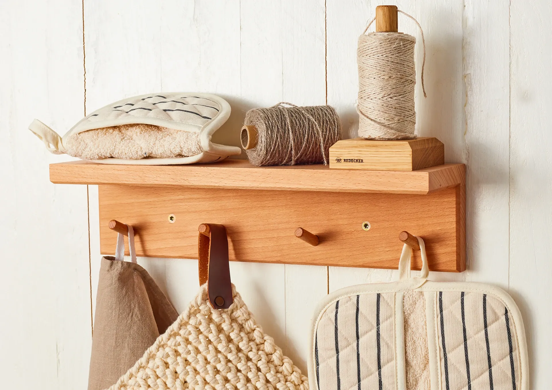 Wooden wall shelf with various items including a spool of twine, a towel, and potholders on a white wooden panel background.