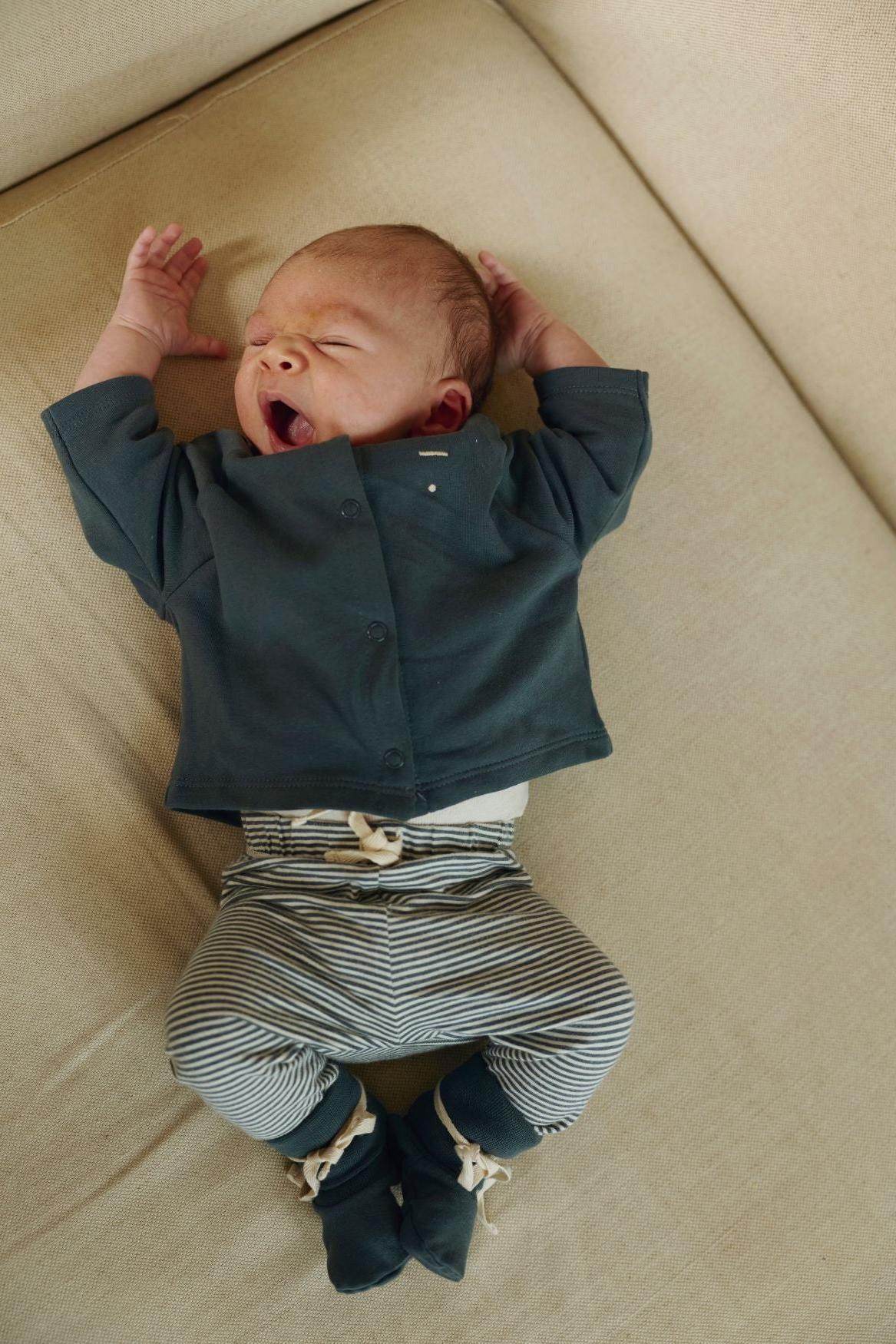 Baby wearing a dark green jacket and striped pants lying on a beige surface.