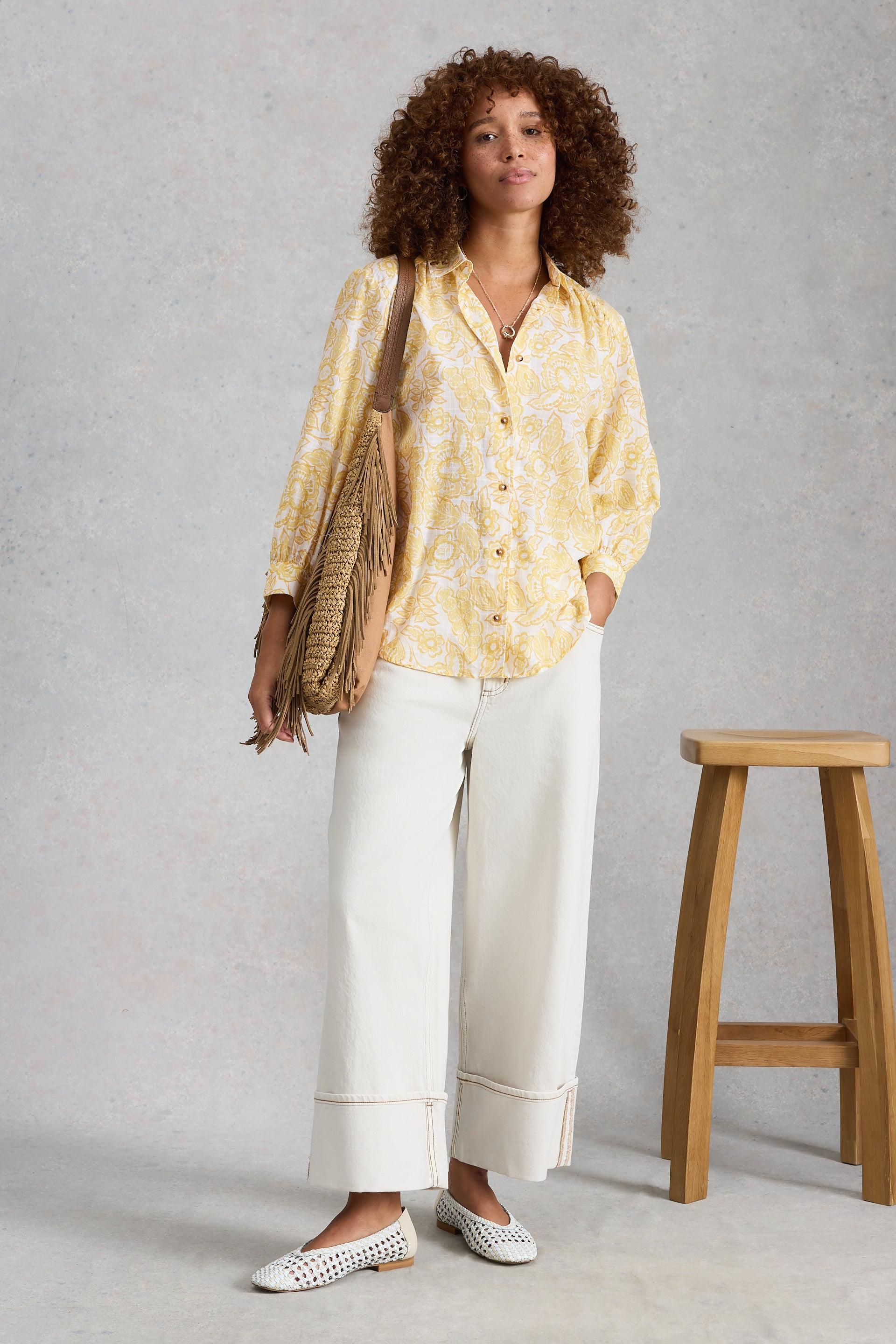 Woman wearing a yellow patterned shirt and white wide leg jeans standing against a gray background